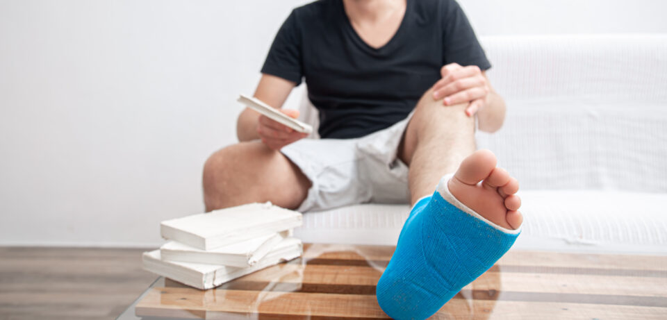 Man with broken leg in blue splint for treatment of injuries from ankle sprain reading books at the home rehabilitation.